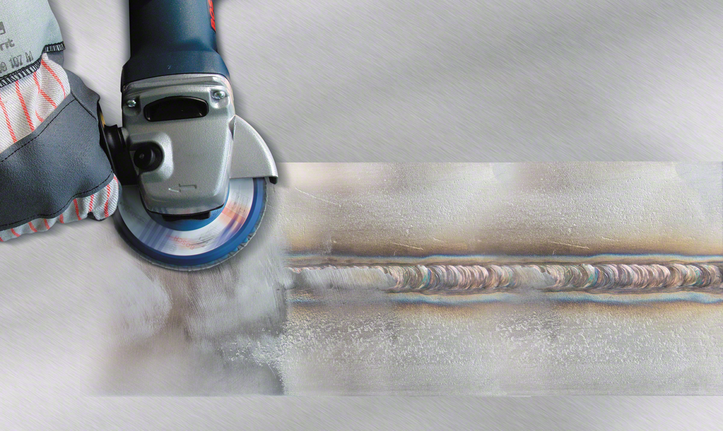 A worker wearing safety equipment grinds a welded seam on metal with an angle grinder.