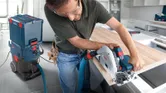 A person wearing safety equipment cuts a wooden board using a circular saw connected to a dust extractor.