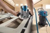 A person guides a router along a board connected to a corded dust extractor.