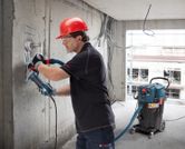 A person wearing safety equipment uses a wall chaser connected to a dust extractor.