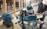 A person wearing safety equipment cuts a wooden beam with a miter saw in a construction area.