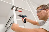 A person wearing safety equipment uses a cordless drill to assemble a cabinet.