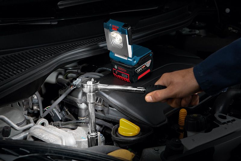 A person works on a car engine using a wrench with a cordless work light nearby.