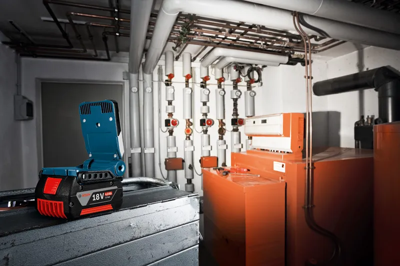GLI VariLED Cordless work light illuminates an industrial boiler room with pipes and orange machinery.