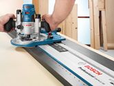 A person guides a wood router along a straight edge track on a light wooden surface.