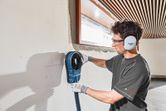 A person wearing safety equipment uses a wall-mounted dust extraction accessory.