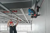 A person wearing safety equipment installs ceiling framing using a laser leveling tool on a wall mount.