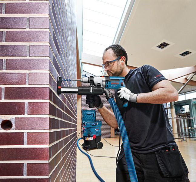 A person wearing safety equipment uses a drill with a dust extraction system on a brick wall.