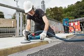 A person wearing safety equipment grinds concrete on a sidewalk with a connected grinder.