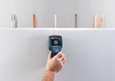 A person uses a wallscanner detector to check behind a drywall surface.