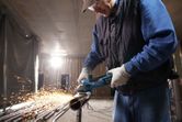 A person wearing safety equipment grinds a metal pipe, producing sparks.