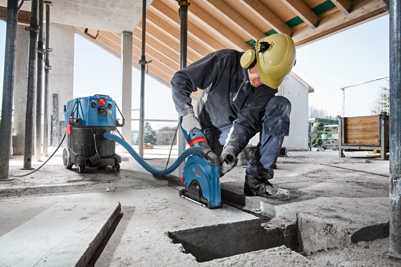A person wearing safety equipment uses a wall chaser with dust extraction on a concrete floor.