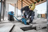 A person wearing safety equipment uses a wall chaser with dust extraction on a concrete floor.
