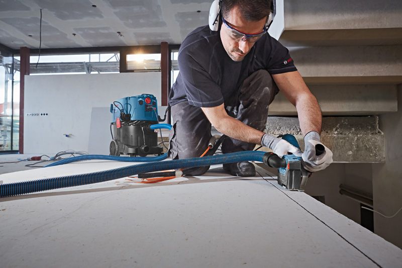 A person wearing safety equipment uses a cutting tool with dust extraction on a drywall panel.
