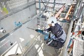 A person wearing safety equipment drills into concrete with a large rotary hammer.