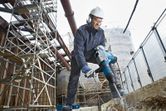 A person wearing safety equipment uses a rotary hammer to break concrete at a construction site.