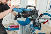 A person wearing safety equipment operates a sliding mitre saw for precise wood cutting.
