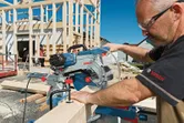 A person wearing safety equipment cuts timber with a sliding mitre saw at a construction site.