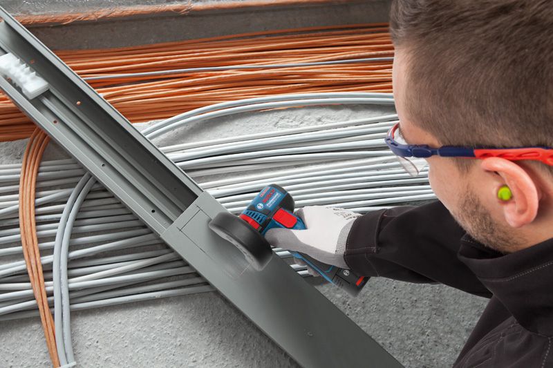 A person wearing safety equipment cuts a metal cable tray with a cordless angle grinder.