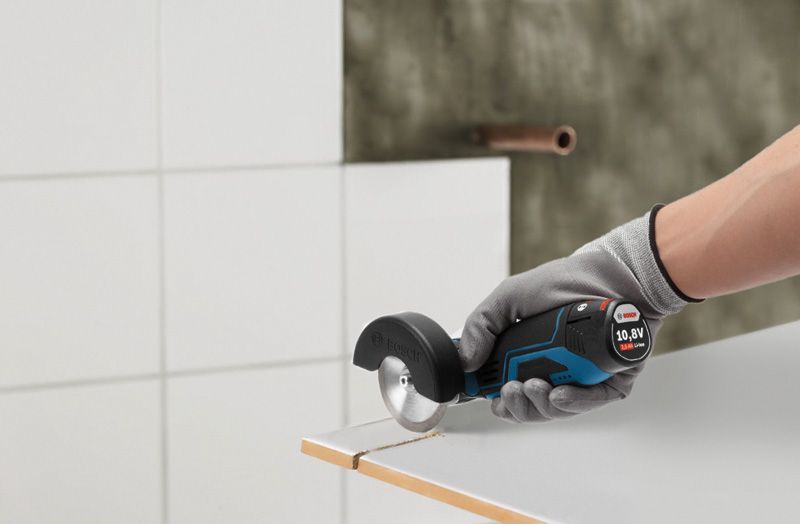 A person wearing safety equipment cuts tile with a small cordless angle grinder.