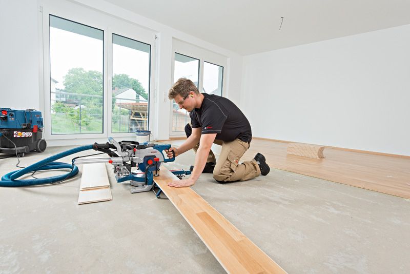 A person wearing safety equipment cuts laminate flooring with a sliding mitre saw.