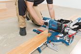 A person wearing safety equipment uses a sliding mitre saw to cut laminate flooring.