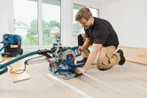 A person wearing safety equipment operates a sliding mitre saw to cut flooring indoors.