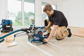 A person wearing safety equipment operates a sliding mitre saw to cut flooring indoors.