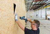 A person wearing safety equipment uses a power saw to cut a rectangular opening in a wall.