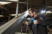 A person wearing safety equipment uses a drill to make a hole in a metal beam.