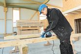A person wearing safety equipment uses a planer to smooth wooden beams indoors.