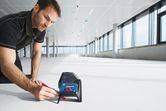 Person marks a line on the floor using a laser leveling tool in a large indoor space.