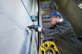A person wearing safety equipment drills into a concrete wall with a rotary hammer.