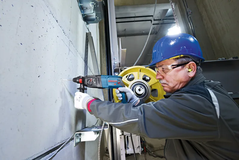 A person wearing safety equipment drills into concrete with a rotary hammer.