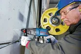 A person wearing safety equipment drills into concrete with a rotary hammer near a yellow pulley.