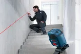 A person wearing safety equipment uses a laser leveling tool to mark a line on a stairwell wall.
