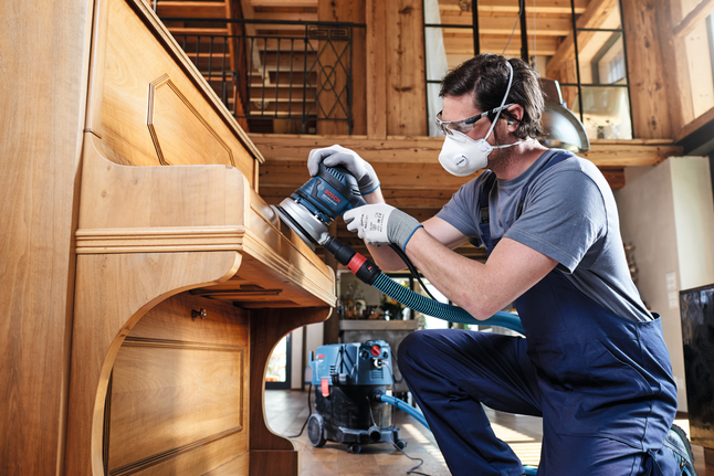 C430 sanding sheet, pack of 5 Person wearing safety equipment sands a wooden piano using a power sander.