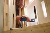 A person wearing safety equipment cuts a wooden board with a cordless multi-tool.