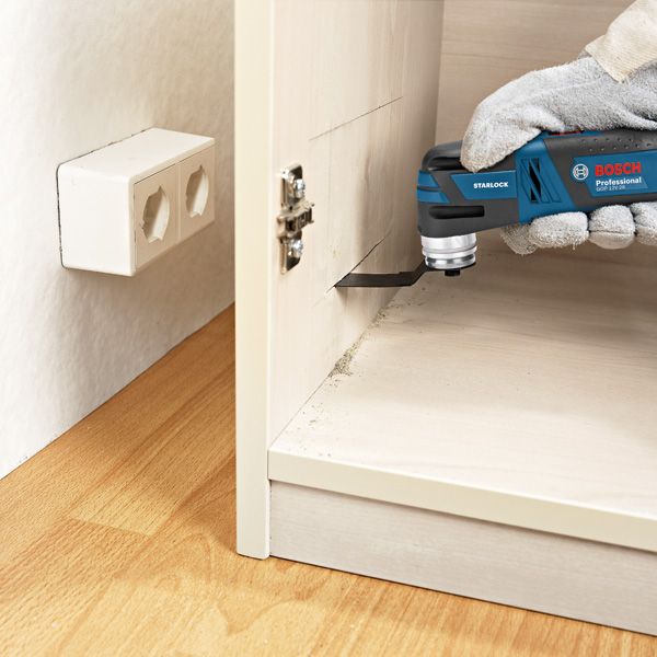 A person wearing safety equipment uses a cordless multi-cutter to cut wood inside a cabinet.