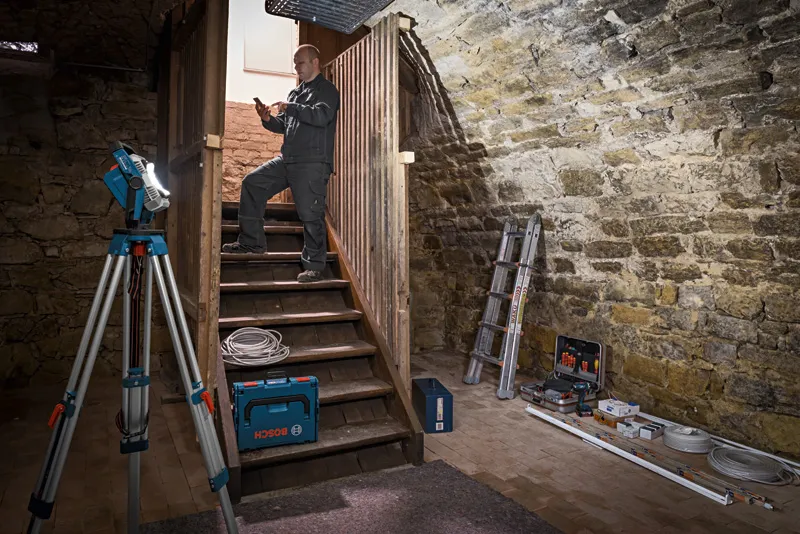 A person wearing safety equipment uses a laser leveling tool on a tripod in a basement.