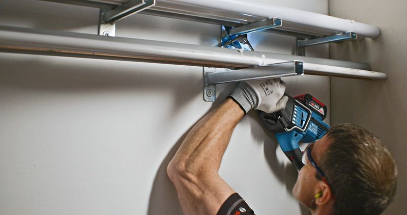 Person wearing safety equipment uses a power drill to mount metal pipes on a wall.
