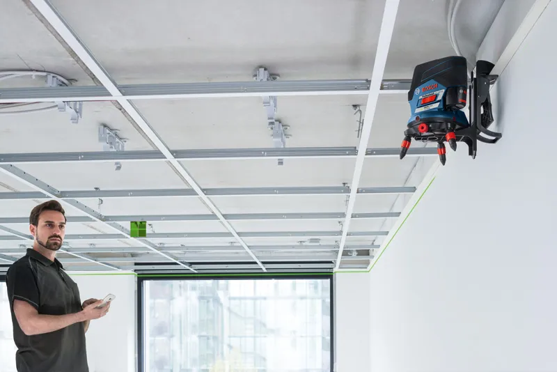 Person using a phone observes a laser leveling tool projecting a green line on a suspended ceiling frame.
