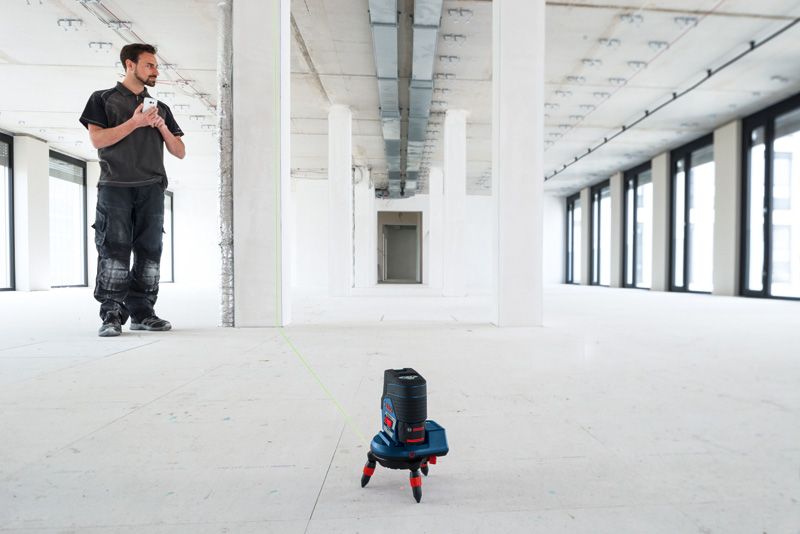 A person stands in a large empty room while a laser leveling tool accessory projects a green line.
