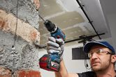 A person wearing safety equipment drills into a brick wall with a cordless drill.