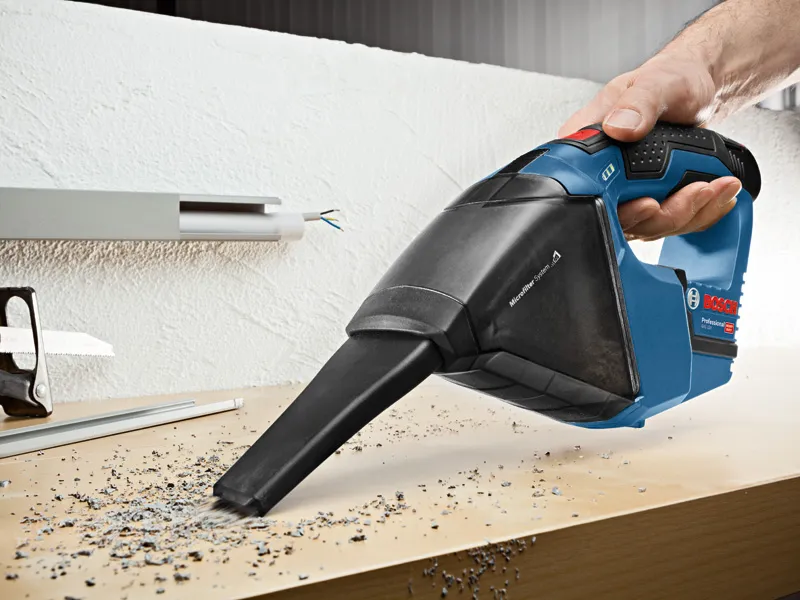 A person uses a cordless vacuum cleaner to pick up debris from a workbench.