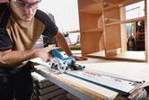 A person wearing safety equipment uses a cordless circular saw to cut wood in a workshop.