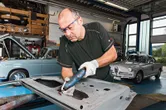 A person wearing safety equipment uses a cordless rotary tool to grind metal in a garage.