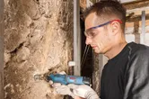 A person wearing safety equipment grinds a wall using a cordless rotary tool.