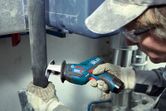 A person wearing safety equipment uses a cordless reciprocating saw to cut a pipe.