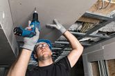 A person wearing safety equipment installs drywall overhead using a cordless screwdriver.